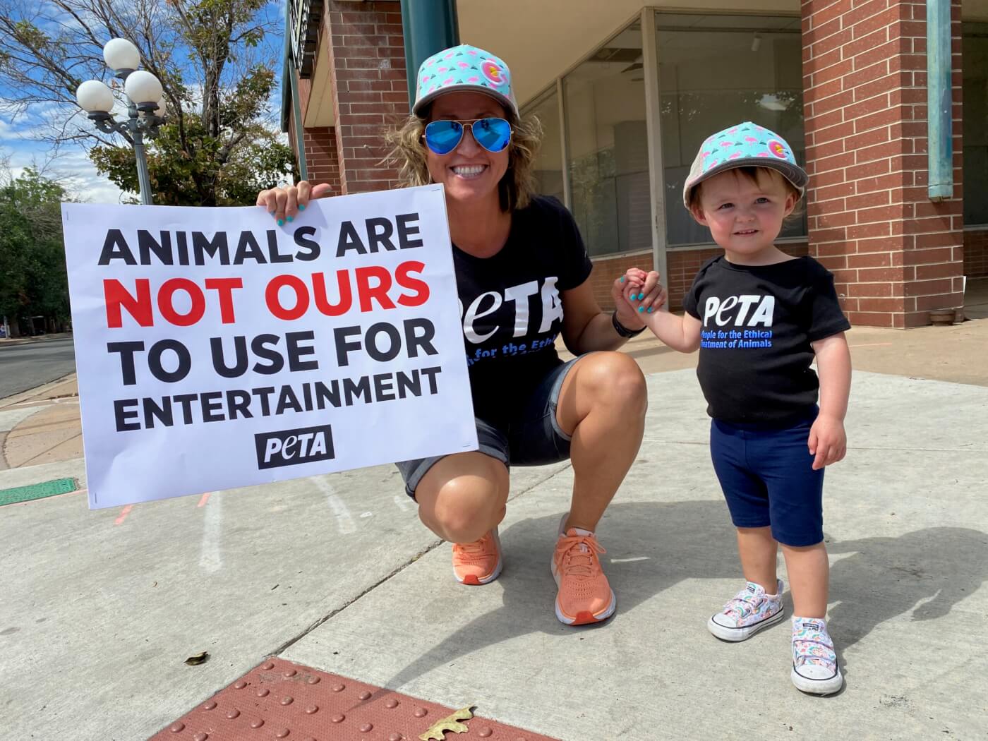 child and her mom on the sidewalk at a PETA protest holding signs that say "Animals are not ours to use for entertainment"