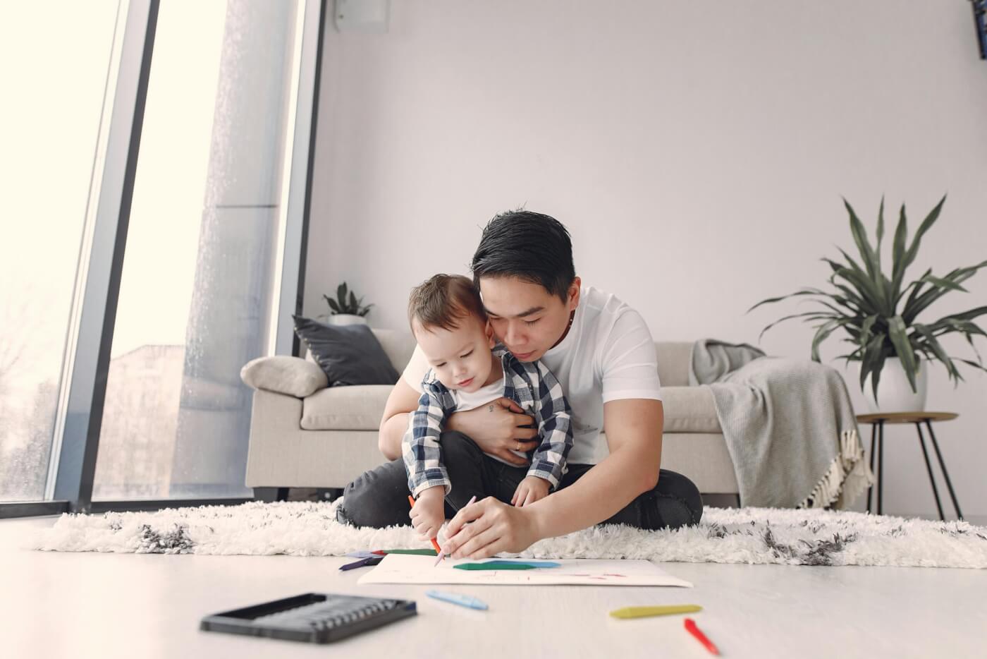 father holding his young toddler on the floor as they do arts and crafts. 