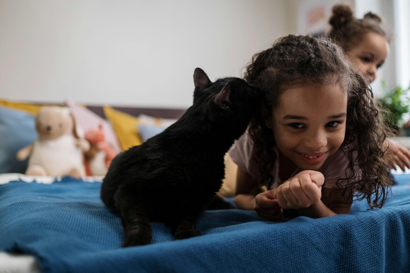 girl is laying on her bed with her younger sister behind her getting cuddles from her black cat.