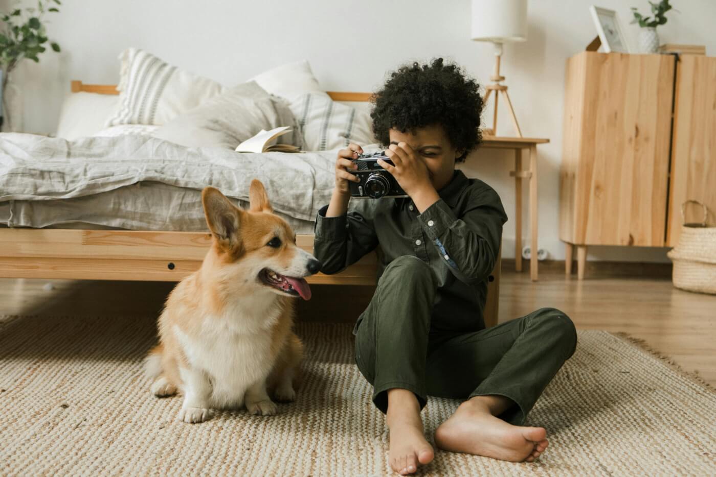 boy sitting on the floor near the foot of his bed in his bedroom taking a photo of his dog.